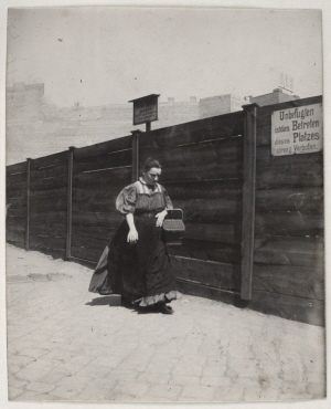 Woman with a basket in front of a site fence, autumn 1898